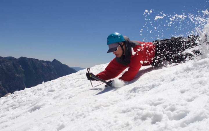 A person wearing mountaineering gear slides down a snowy incline on their belly.
